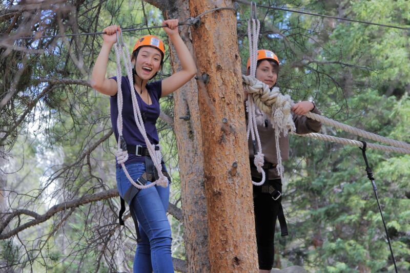 Two young women are enjoying a ropes course in a forest setting. Both are wearing helmets and safety harnesses, indicating they are engaged in a safe, adventurous activity. The woman on the left is smiling broadly and holding onto a rope, while the other woman is standing on a platform attached to a tree. The background features lush green trees, suggesting a natural and scenic environment for the ropes course.